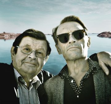 Kaksi miestä poseeraa rauhallisen järven rannalla, jonka taustalla on kallioisia saaria. Toinen mies, vanhempi ja silmälasipäinen, hymyilee lämpimästi. Toisella, nuoremmalla, on aurinkolasit ja hänellä on käsi ensimmäisen miehen olkapään ympärillä.