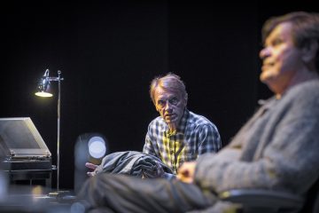 Kaksi miestä istuu hämärästi valaistulla näyttämöllä; toinen, jolla on ruudullinen paita, katsoo keskittyneesti eteenpäin, takki kädessään, kun taas toinen on epäselvästi etualalla. Pöytälamppu valaisee kohtausta.