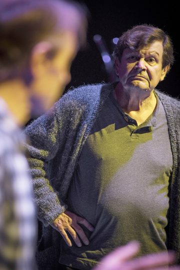 Vanhempi mies, jolla on vakava ilme, seisoo toinen käsi lantiollaan, päällään harmaa neuletakki ja paita, ja katsoo etualalla olevaa epäselvää henkilöä. Tausta on tumma.