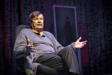 Vanhempi mies istuu tuolilla lavalla yllättynyt ilme kasvoillaan, kädet ojennettuina. Hänellä on harmaa pusero ja paita, ja hänen takanaan on tumma tausta ja kehystetty kuva.