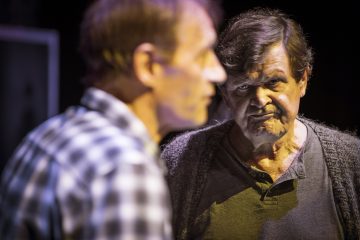 Vanhempi mies, jolla on harmaat hiukset ja vakava ilme, katsoo etualalla olevaa toista miestä, joka on epätarkka ja jolla on ruudullinen paita. Kohtaus vaikuttaa jännittyneeltä ja dramaattiselta, mahdollisesti näyttämöesityksestä.