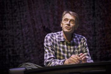 Vanhempi vaaleanruskeatukkainen mies, jolla on ruudullinen paita, istuu pöydän ääressä kädet ristissä ja katsoo hieman ylöspäin tummaa, kuvioitua taustaa vasten.