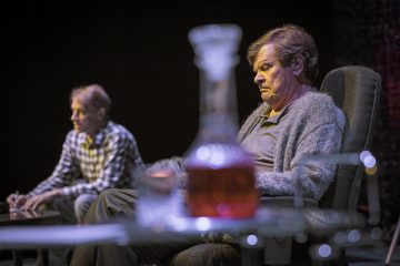 Kaksi miestä istuu sisätiloissa; toinen etualalla näyttää mietteliäältä harmaassa villapaidassaan, kun taas toinen, ruudulliseen paitaan pukeutunut mies on hämärtynyt taustalla. Heidän edessään pöydällä on lasinen dekantteri, jossa on meripihkanväristä nestettä.