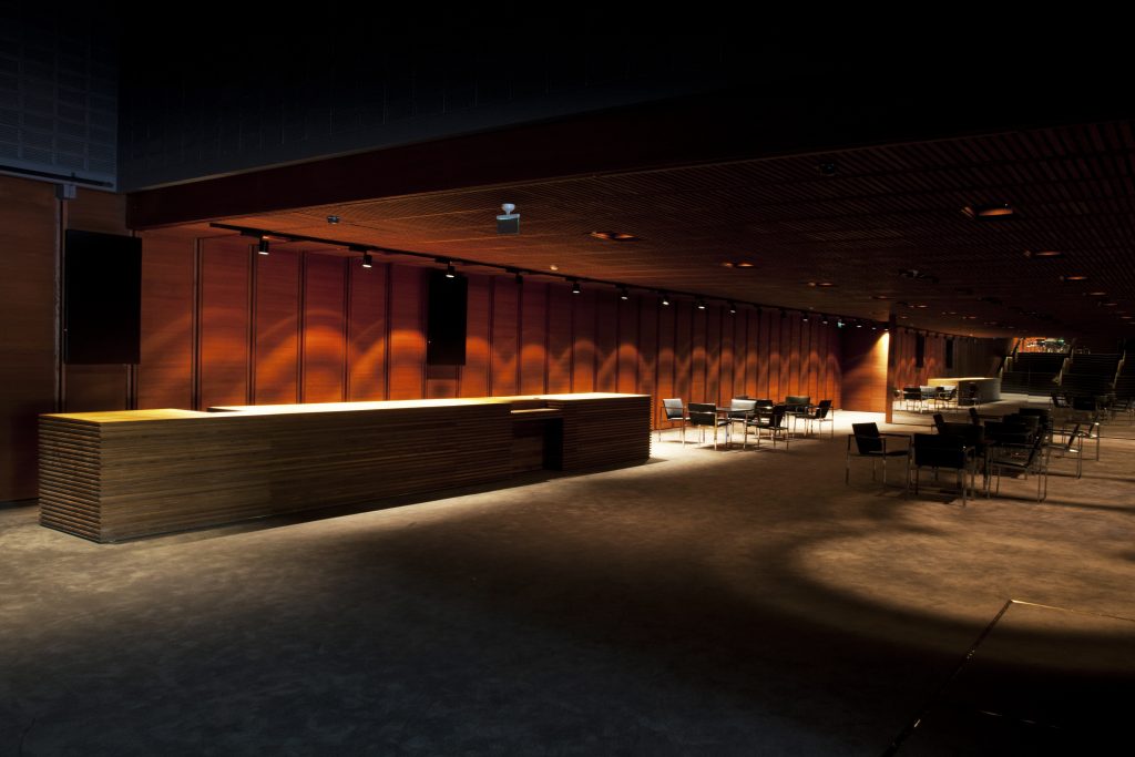 Moderni, hämärästi valaistu baari, jossa on pitkä puinen tiski ja hajallaan olevia mustia tuoleja ja pöytiä; lämmin valo heittää varjoja puuseinille luoden viihtyisän, minimalistisen ilmapiirin.