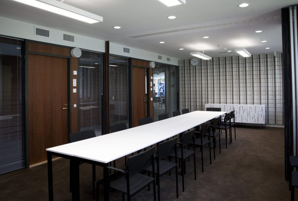 Moderni kokoushuone, jossa on pitkä valkoinen pöytä, jota ympäröivät mustat tuolit, lasiseinät, puuovet, seinäkello ja kattovalaisimet. Huone vaikuttaa tyhjältä ja hyvin valaistulta.