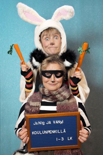 Kaksi eläinasuihin pukeutunutta ihmistä poseeraa leikkisästi; toinen on pukeutunut jänikseksi, jolla on korvat ja porkkanat, ja istuu toisen raidalliseen paitaan ja naamariin pukeutuneen henkilön takana pitäen kädessään suomenkielistä kylttiä, jossa lukee Jänis ja Vanki koulunpenkillä 1-3 lk.