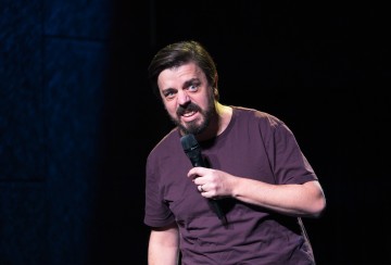 Mies, jolla on parta ja viikset ja jolla on ruskeankeltainen T-paita, pitää mikrofonia ja esiintyy lavalla tummalla taustalla, vaikuttaen eloisalta ja ilmeikkäältä.