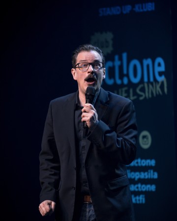 Mies, jolla on silmälasit, viikset ja pukinparta, esittää stand up -komiikkaa lavalla mikrofoni kädessään. Hänellä on yllään musta bleiseri ja paita. Taustalla näkyy sinisellä valaistu tapahtumabanneri ja teksti.