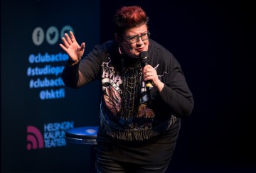 Lyhyet tummat hiukset ja punaiset raidat omaava esiintyjä, jolla on silmälasit ja musta graafinen paita, elehtii elävästi mikrofoni kädessään Helsingin kaupunginteatterin lavalla.