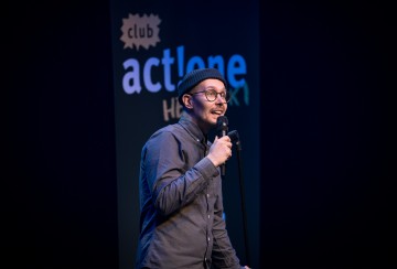 Henkilö, jolla on silmälasit, pipo ja napillinen paita, pitää mikrofonia esiintyessään lavalla. Heidän takanaan olevassa kyltissä lukee club actlone. Lava on hämärästi valaistu ja sen tausta on tumma.