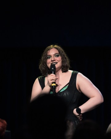 Lyhyet ruskeat hiukset omaava ja rohkeasti meikattu henkilö pitelee mikrofonia ja hymyilee esiintyessään lavalla hihattomassa mustassa asussa. Tausta on tumma, ja etualalla näkyy yleisö.