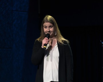 Nainen, jolla on pitkät vaaleat hiukset, musta neuletakki ja valkoinen toppi, seisoo lavalla mikrofoni kädessään ja puhuu vakavalla ilmeellä tummaa taustaa vasten.