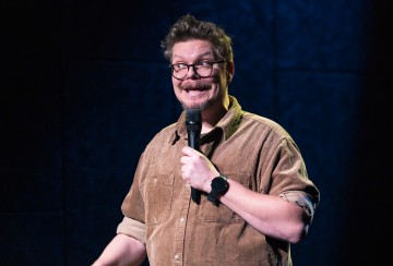 Silmälasipäinen ja ruskeaan paitaan pukeutunut henkilö pitelee mikrofonia, hymyilee leveästi tummalla lavalla ja näyttää esittävän stand up -komiikkaa.