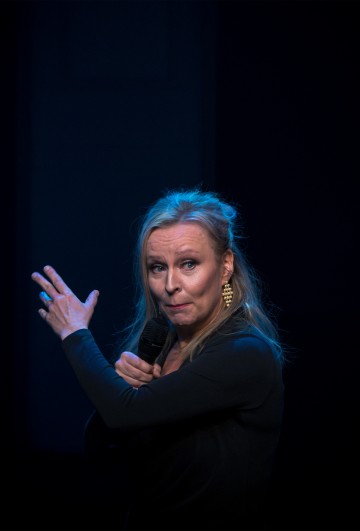 Vaaleatukkainen nainen pitää mikrofonia ja elehtii vasemmalla kädellään esiintyessään lavalla hämärässä valaistuksessa. Hänellä on yllään kultaiset korvakorut ja musta asu.