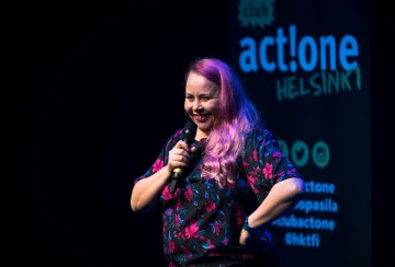 Pitkät vaaleanpunaiset hiukset omaava nainen pitelee mikrofonia ja hymyilee lavalla. Hän seisoo mustan taustakuvan edessä, jossa lukee