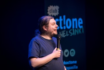 Henkilö, jolla on pitkät hiukset poninhännässä, pitää mikrofonia ja puhuu lavalla. Heidän takanaan on kyltti, jossa on teksti club artlone HELSINKI ja sosiaalisen median kuvakkeita. Kohtaus on kirkkaasti valaistu tummaa taustaa vasten.