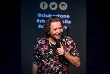 Lavalla seisoo hymyillen ja mikrofoni kädessään pitkät hiukset ja partainen mies, jolla on musta kukkapaita. Hänen takanaan olevalla taululla näkyy sosiaalisen median kahvoja ja hashtageja.