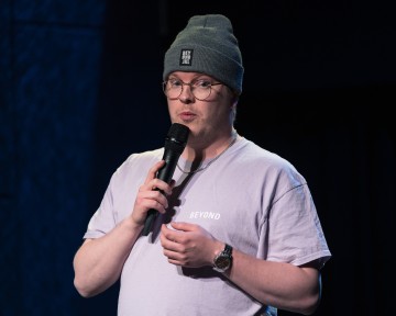 Henkilö, jolla on silmälasit, harmaa pipo ja vaaleanvioletti t-paita, pitelee mikrofonia puhuessaan lavalla tummansinisellä taustalla.