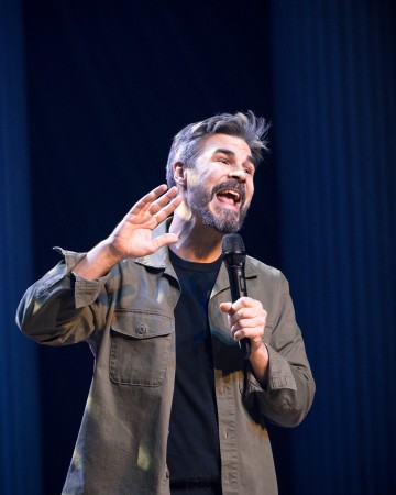 Harmaahiuksinen ja parrakas mies, jolla on toisessa kädessä mikrofoni, elehtii toisella kädellä korvansa lähellä puhuessaan tai esiintyessään lavalla. Hänellä on yllään vihreä takki mustan paidan päällä.