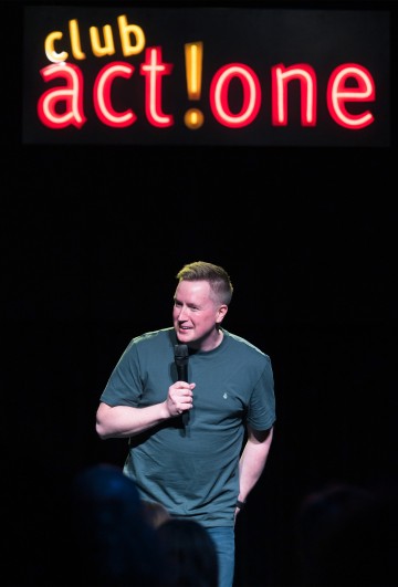 Vihreään t-paitaan pukeutunut henkilö pitelee mikrofonia ja esiintyy lavalla kirkkaan kyltin alla, jossa lukee club act! one. Tausta on tumma, ja yleisö näkyy etualalla.