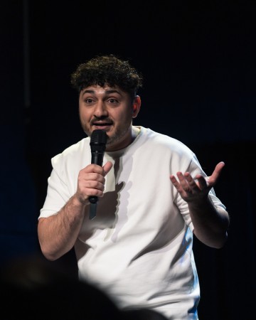 Kiharatukkainen, parrakas, valkoiseen paitaan pukeutunut mies pitelee mikrofonia ja elehtii toisella kädellään puhuessaan lavalla hämärässä valaistussa ympäristössä.