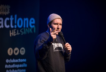 Pipoon ja tummaan collegepaitaan pukeutunut henkilö pitelee mikrofonia ja elehtii esiintyessään lavalla. Taustalla näkyy sininen ja musta kyltti, jossa on sosiaalisen median kädensijat ja act!one HELSINKI -teksti.