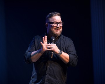 Silmälasipäinen ja parrakas mies, jolla on musta paita, pitää mikrofonia ja hymyilee esiintyessään lavalla tummalla taustalla.