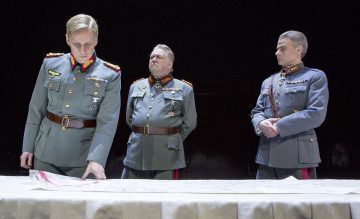 Kolme sotilaspukuista miestä seisoo pöydän ympärillä, jossa on suuri kartta; yksi tutkii karttaa, kun muut katsovat vakavasti, ja kaikki vaikuttavat korkea-arvoisilta upseereilta dramaattisessa, hämärästi valaistussa ympäristössä.