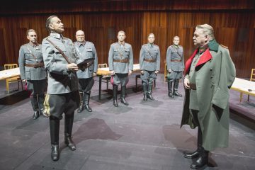Kuusi sotilasunivormuihin pukeutunutta miestä seisoo lavalla puupaneloidussa huoneessa. Viisi miestä seisoo varuillaan, kun taas yksi heistä kohtaa toisen pitkätakkisen miehen, mikä viittaa teatteriesityksen dramaattiseen tai vastakkainasetteluun liittyvään hetkeen.