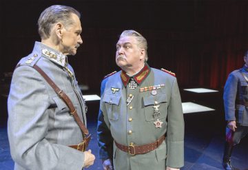 Kaksi miestä, joilla on koristellut sotilasasut, kohtaavat toisensa näyttämöllä intensiivisesti, mikä viittaa dramaattiseen yhteenottoon. Taustalla on pimeä, punaiset verhot ja valaistut lavasteet. Toinen hahmo seisoo osittain näkyvissä oikealla.