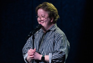 Silmälasipäinen ja raitapaitainen henkilö seisoo mikrofonin edessä hymyillen ja kädet yhteen lyötynä tummaa taustaa vasten.