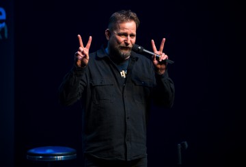 Parrakas mies seisoo lavalla mikrofoni toisessa kädessä ja tekee rauhanmerkkejä molemmin käsin. Hänellä on tumma paita ja hän seisoo tumman taustan edessä, jonka lähellä on jakkara.