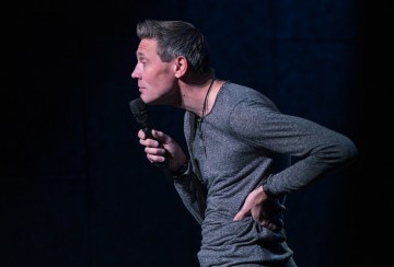 Lyhythiuksinen henkilö pitää mikrofonia toisessa kädessään ja nojaa eteenpäin käsi alaselällään ja näyttää esittävän stand up -komiikkaa hämärästi valaistulla lavalla.