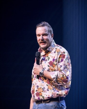 Lyhythiuksinen, parrakas mies, jolla on kukkapaita ja farkut, pitää mikrofonia ja puhuu lavalla tummaa, pehmeästi valaistua taustaa vasten.