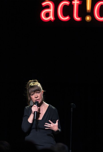 Mustiin pukeutunut nainen pitelee mikrofonia ja elehtii esiintyessään lavalla. Hänen takanaan on neonkyltti, jossa lukee act! tummaa taustaa vasten.