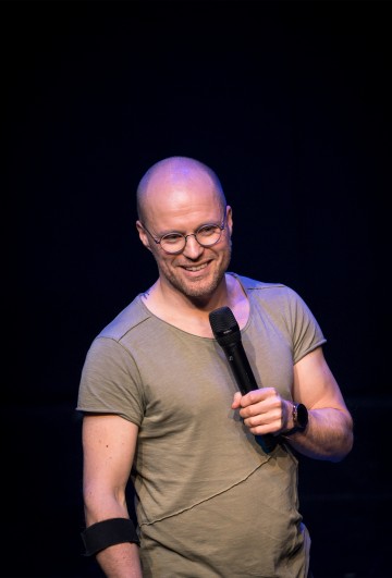 Hymyilevä kalju mies, jolla on silmälasit, vaaleanvihreä t-paita ja älykello, pitelee mikrofonia esiintyessään lavalla tummaa taustaa vasten.