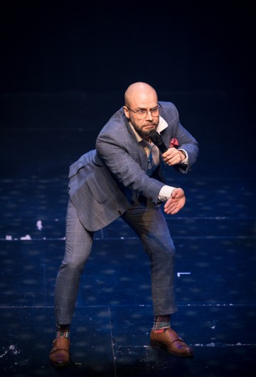 Harmaaruutuiseen pukuun ja ruskeisiin kenkiin pukeutunut kalju mies pitää mikrofonia, nojaa eteenpäin ja tekee eleitä pimeällä lavalla.