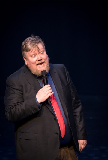 Parrakas mies, jolla on puku ja punainen solmio, pitää mikrofonia ja puhuu vilkkaasti lavalla tummaa taustaa vasten.