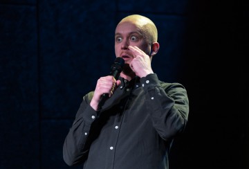 Tummapaitainen kalju mies pitää mikrofonia kädessään ja näyttää yllättyneeltä. Hän seisoo hämärästi valaistulla näyttämöllä, silmät suurina ja toinen käsi kasvojaan koskettaen.