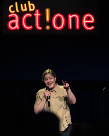 Mikrofonia pitelevä henkilö hymyilee ja osoittaa lavalla rauhanmerkkiä kirkkain kirjaimin kirjoitetun club act! one -kyltin alla.