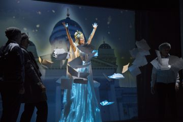 Nainen, jolla on hohtavan sininen puku ja kruunu, seisoo kädet kohotettuna, kun paperit lentävät hänen ympärillään, kolme ihmistä seisoo lähellä ja taustalla näkyy suuri rakennus.
