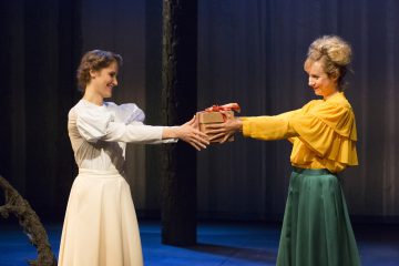 Lavalla seisoo kaksi värikkäisiin pukuihin pukeutunutta naista. Toinen, valkoiseen puseroon ja hameeseen pukeutunut, vastaanottaa käärityn lahjan toiselta, joka on pukeutunut keltaiseen puseroon ja vihreään hameeseen. Molemmat hymyilevät, ja heidän takanaan on tumma teatteritausta.