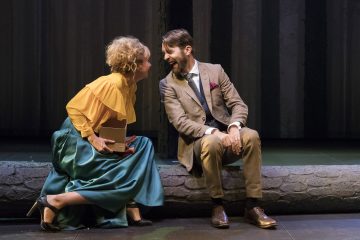 Keltaiseen puseroon ja vihreään hameeseen pukeutunut nainen istuu ruskeaan pukuun pukeutuneen miehen vieressä tukin päällä, molemmat nauravat ja nojaavat toisiinsa; naisella on kirja sylissään, ja näyttämön tausta on hämärästi valaistu.