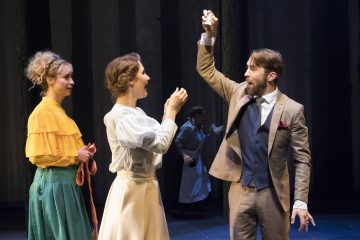 Lavalla esiintyy kolme näyttelijää; pukumies nostaa innostuneesti kätensä kädessään jotain, ja kaksi aikakauden pukuihin pukeutunutta naista, joista toinen on keltainen ja vihreä ja toinen valkoinen, katselee häntä. Heidän takanaan on hämärästi valaistu metsätausta.