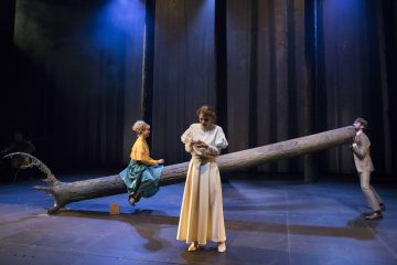 Lavalla on kolme näyttelijää, jotka esiintyvät suuren kaatuneen puun vieressä; yksi keltaiseen ja tiilenpunaiseen pukeutunut nainen istuu puun rungolla, toinen valkoisiin pukeutunut nainen seisoo ja lukee, kun taas harmaapukuinen mies tukee puun päätä. Tausta muistuttaa tummaa metsää.