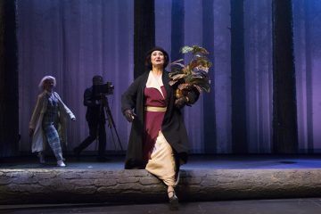 Pitkään takkiin ja hattuun pukeutunut nainen pitelee ruukkukasvia seisoessaan kaatuneen tukin päällä näyttämöllä, taustalla kaksi ihmistä ja kamera korkeiden puiden taustaa vasten.