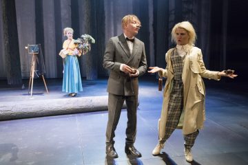 Kolme näyttelijää esiintyy näyttämöllä metsäisessä ympäristössä; yksi puvussa, yksi ruudullisessa asussa ja kevyessä takissa elehtien ilmeikkäästi, ja yksi sinisessä hameessa vanhan kameran ja puiden rekvisiitan lähellä taustalla.