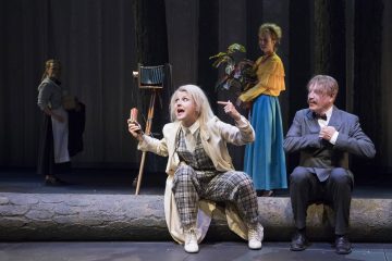 Ruudulliseen pukuun ja valkoiseen takkiin pukeutunut nainen istuu vilkkaasti hirren päällä esine kädessään, ja hänen vieressään istuu pukuun pukeutunut mies. Taustalla seisoo kaksi naista vintage-kameran ja ruukkukasvin vieressä metsäisessä ympäristössä.