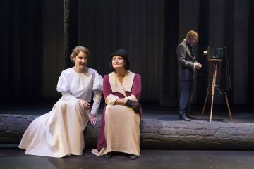 Kaksi aikakauden asuihin pukeutunutta naista istuu matalalla kivimuurilla, toinen valkoisessa mekossa ja toinen ruskeanruskeassa ja beigen värisessä asussa, jossa on hattu. Heidän takanaan seisoo pukuun pukeutunut mies, joka säätää jalustalla olevaa vintage-kameraa.