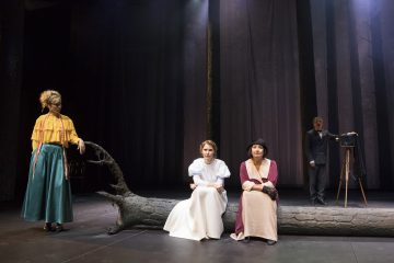 Neljä näyttelijää hämärästi valaistulla näyttämöllä; kaksi naista istuu kaatuneella tukilla, toisella valkoinen mekko, toisella vaaleanpunainen mekko ja hattu. Keltaiseen puseroon pukeutunut nainen seisoo, ja pukuun pukeutunut mies seisoo vanhan kameran vieressä. Tausta on tumma ja metsäinen.